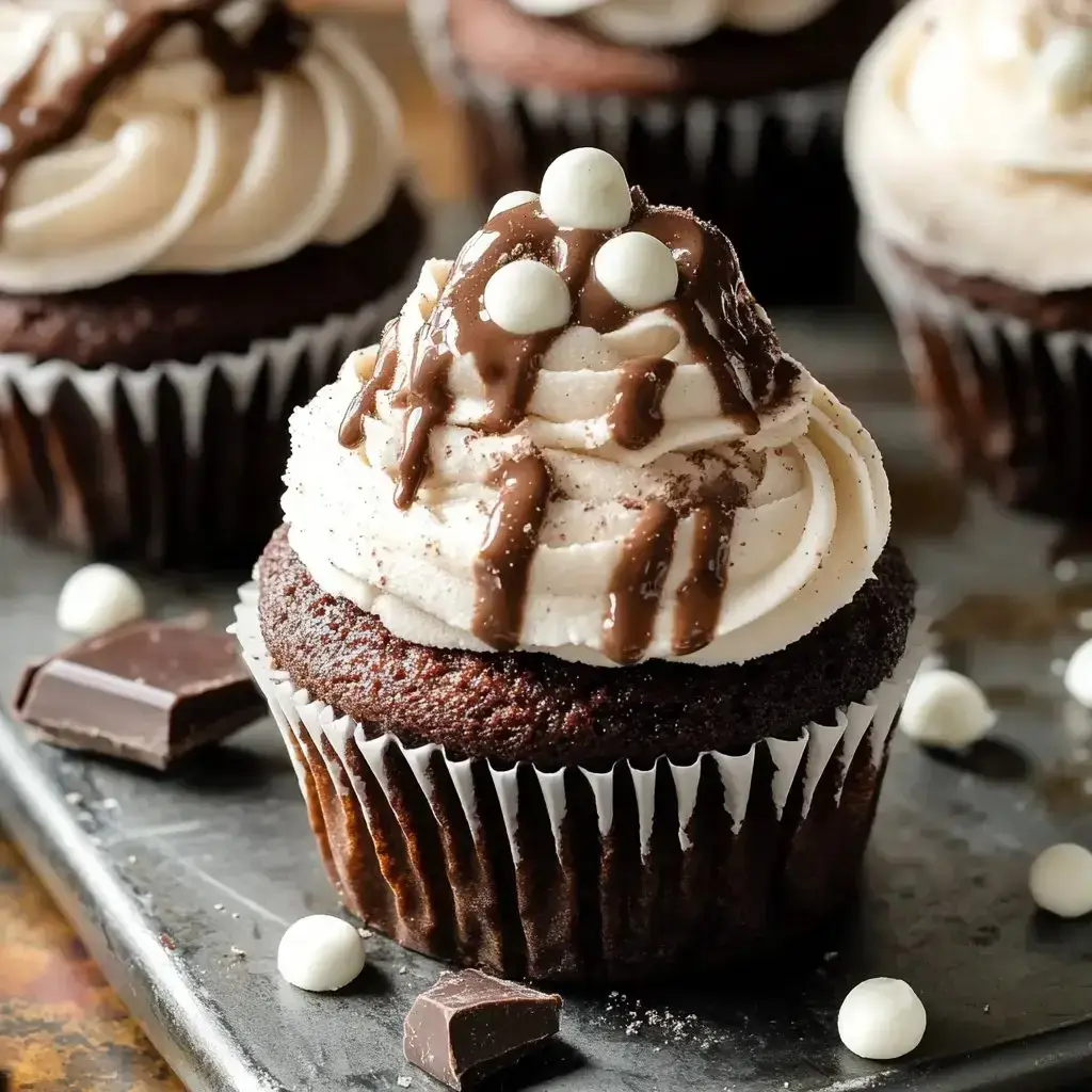 Up-close view of a chocolate cupcake with whipped frosting, chocolate drips, and marshmallows, sitting among more cupcakes and chocolate chunks on a dark table.