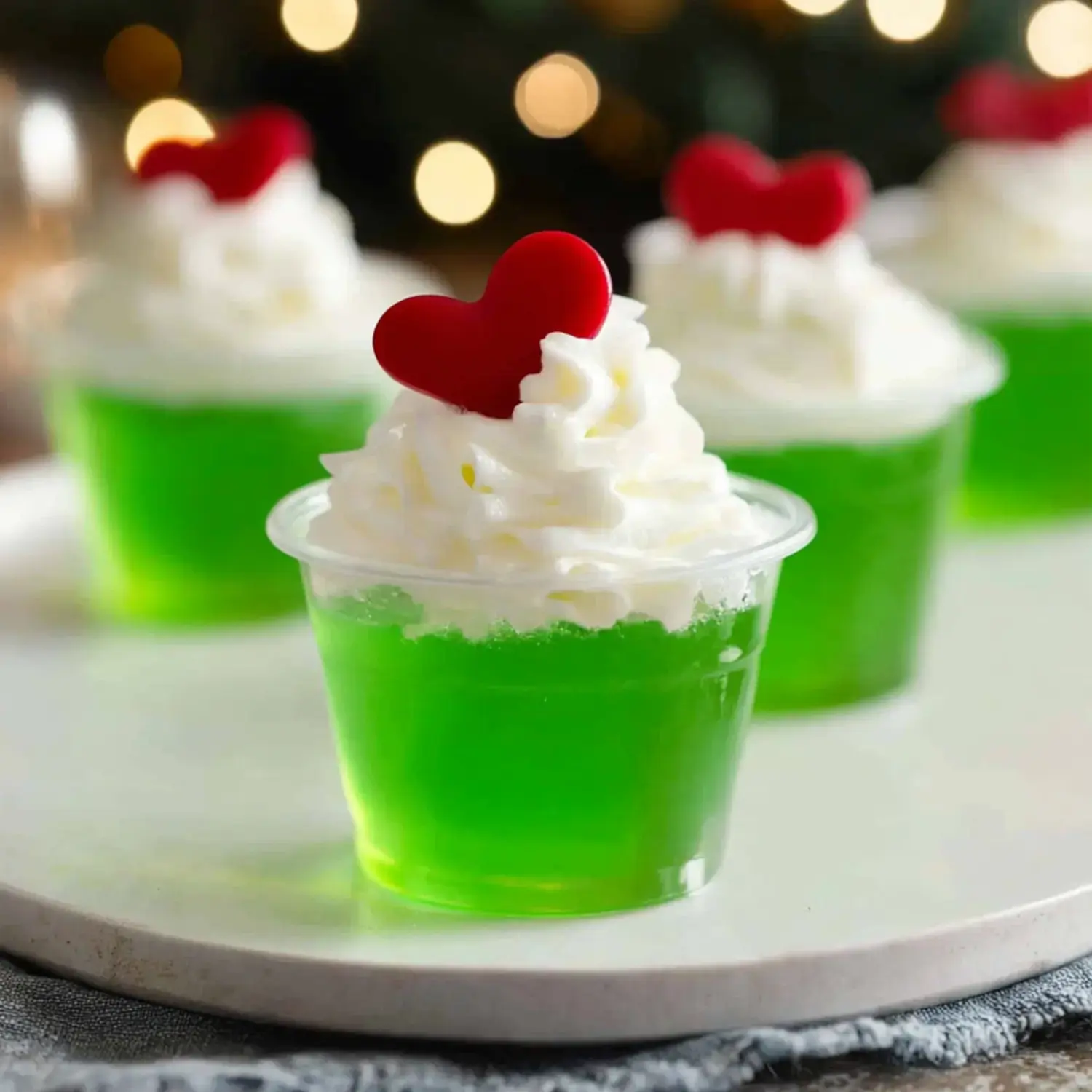 Close-up of green gelatin desserts topped with cream and small red heart sprinkles, with soft festive lights in the background.